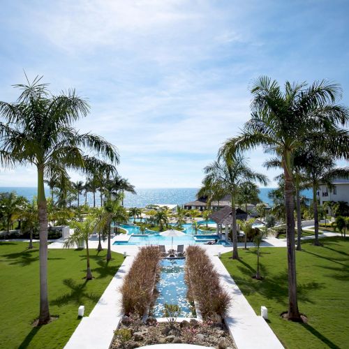 A tropical resort with palm trees, a central water feature, manicured lawns, and poolside buildings overlooking the sea, feeling sunny and serene.