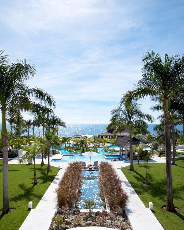 A tropical resort with palm trees, a central water feature, manicured lawns, and poolside buildings overlooking the sea, feeling sunny and serene.