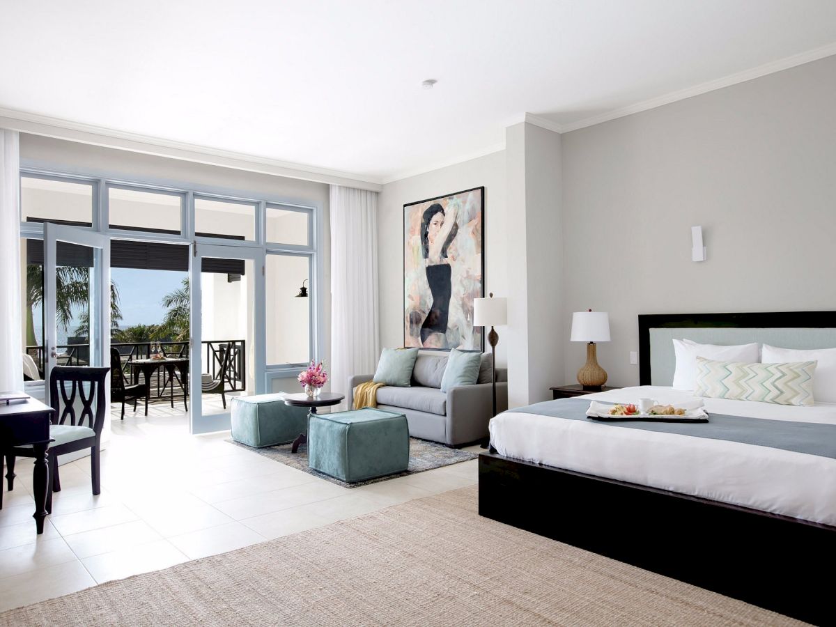 A bright hotel suite with a large bed, gray accent wall, a seating area, and glass doors opening to a balcony with outdoor views.