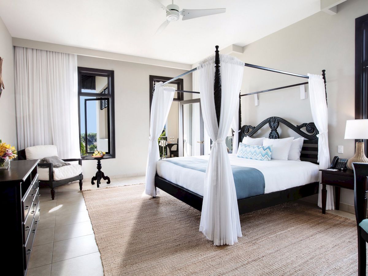 A luxurious four-poster bed with white drapes sits in a bright hotel room, flanked by nightstands, a desk, and seating by windows.