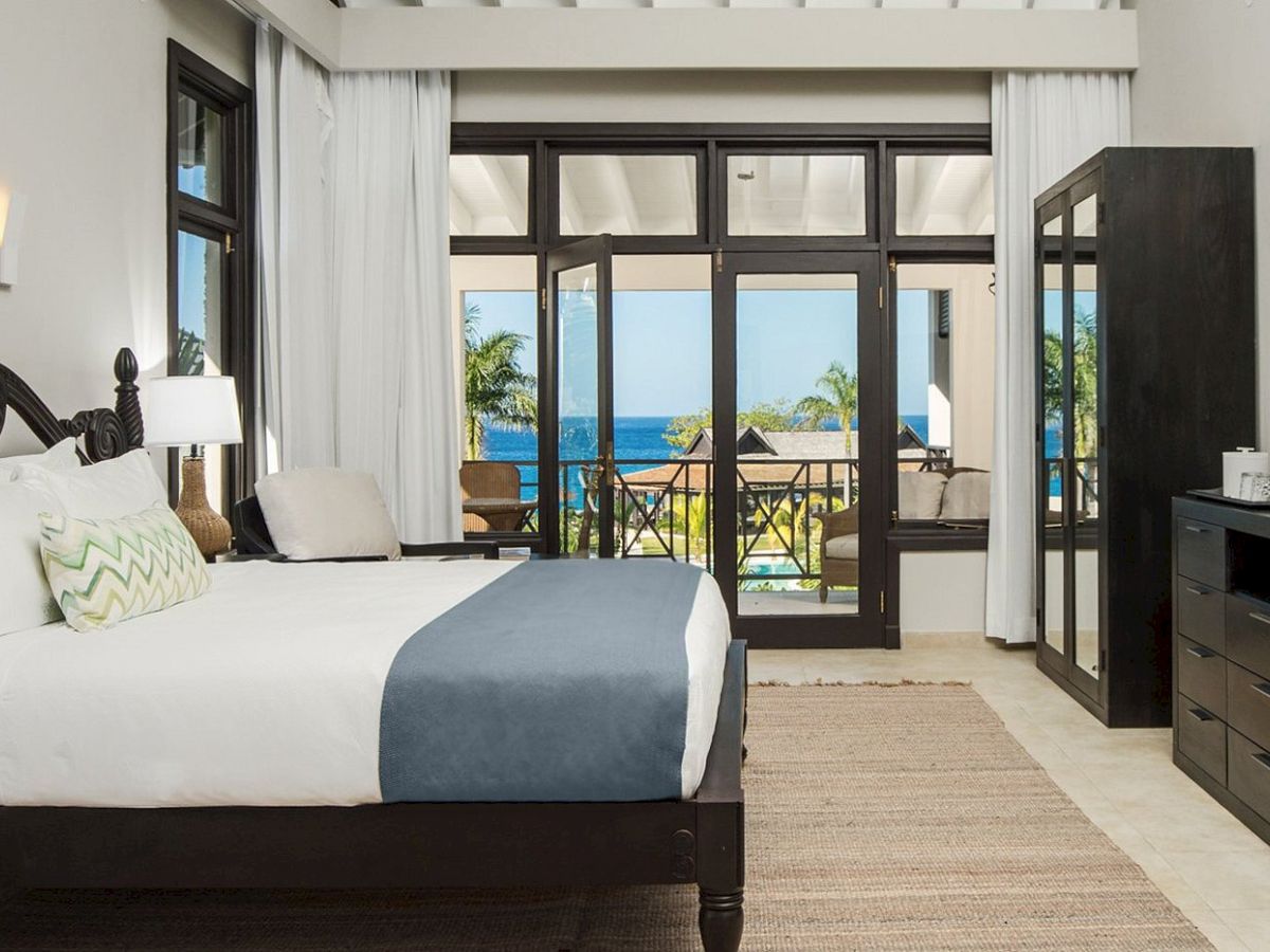 A hotel room with a dark wooden bed, white and blue bedding, a rug, and a balcony view of palm trees and the ocean through glass doors. Ending with a period.