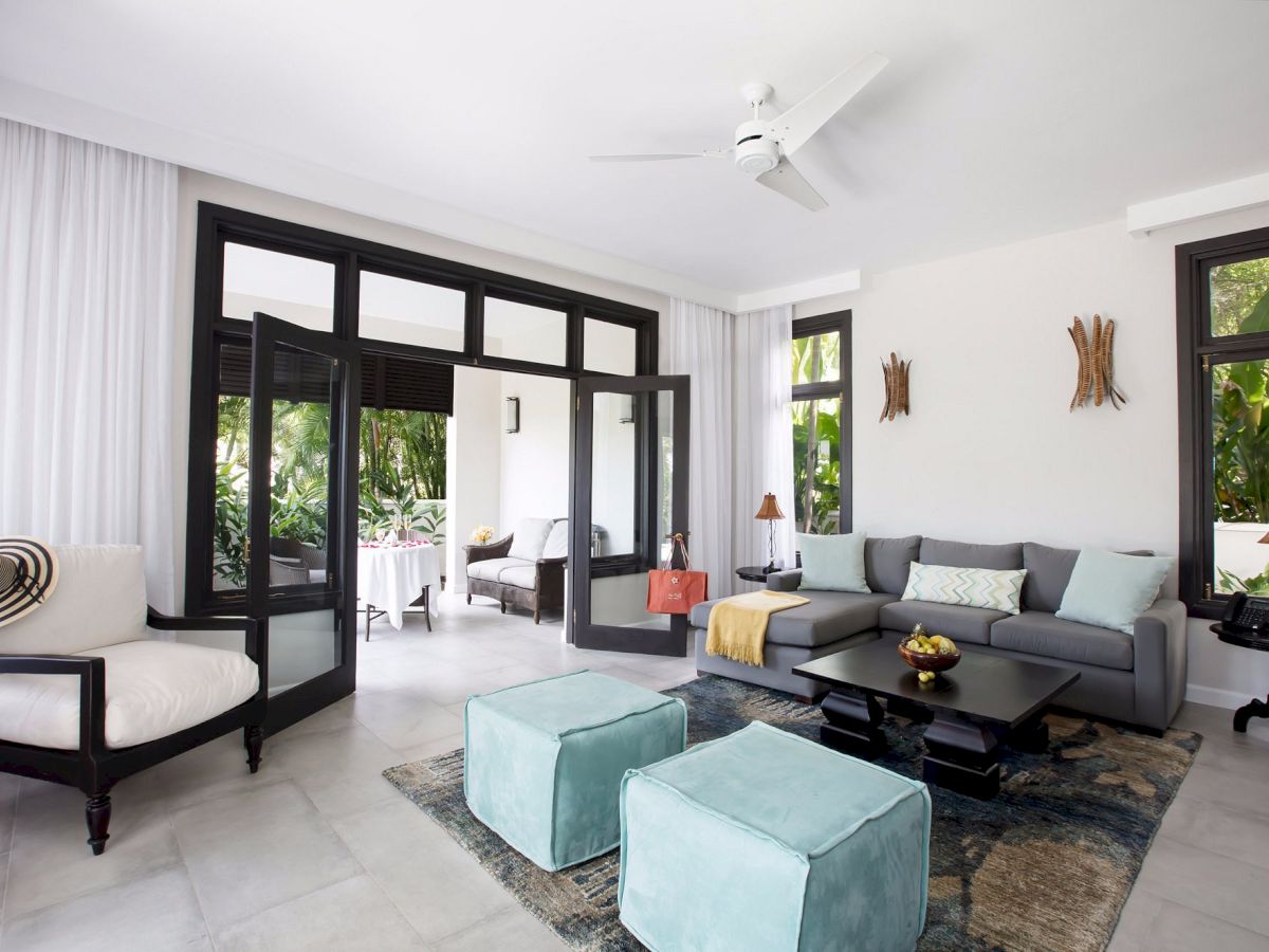 A bright, airy living room with a gray sofa, two turquoise ottomans, a glass coffee table, and large windows opening to a patio, sunlit and stylish.
