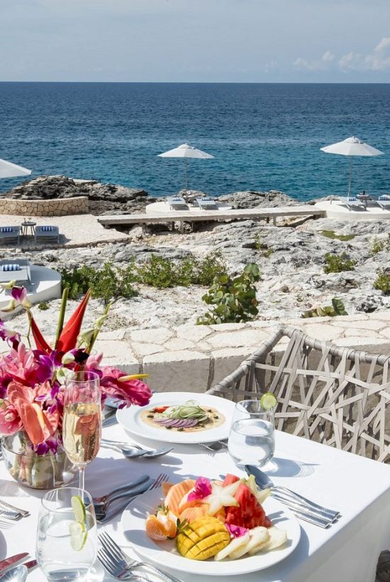 A seaside patio dining scene with a white table set for four, fresh flowers, plates of food, wine glasses, and a stone terrace overlooking the blue ocean under a sunny sky.