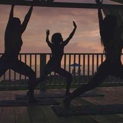 People doing yoga or exercise silhouettes on a wooden deck at sunset, arms raised in celebration, railing and distant horizon in view.