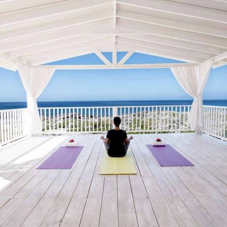 A person sits on a yellow yoga mat under a white gazebo, facing the ocean. Two purple mats flank them on a sunny, open deck.