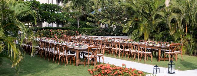 A long row of wooden benches is arranged under a roofed shelter beside a grassy yard, with trees and a white building in the background.