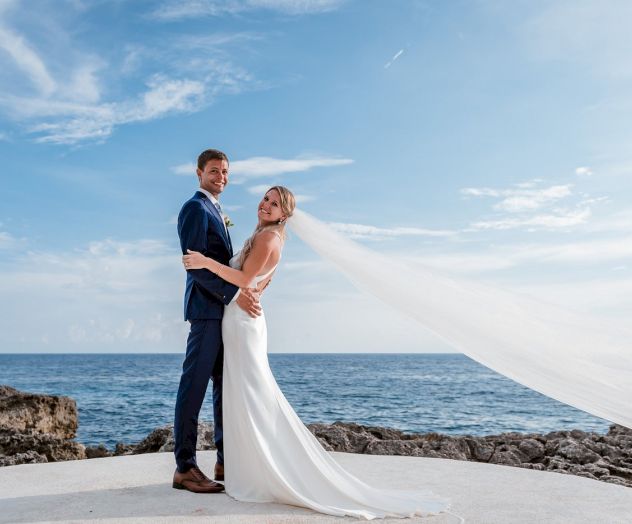 A bride and groom kiss on the beach, holding each other in wedding attire with a sunny, blue sky and calm sea behind them.