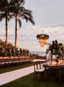 Outdoor wedding reception setup with long tables, string lights, palm trees, and a dramatic hanging flame lantern adorning the aisle, creating a warm festive vibe.