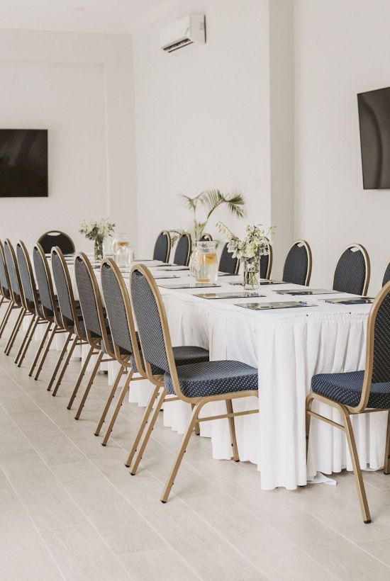 A bright, minimalist conference room with a long table, many chairs, plants, and wall-mounted screens for meetings.
