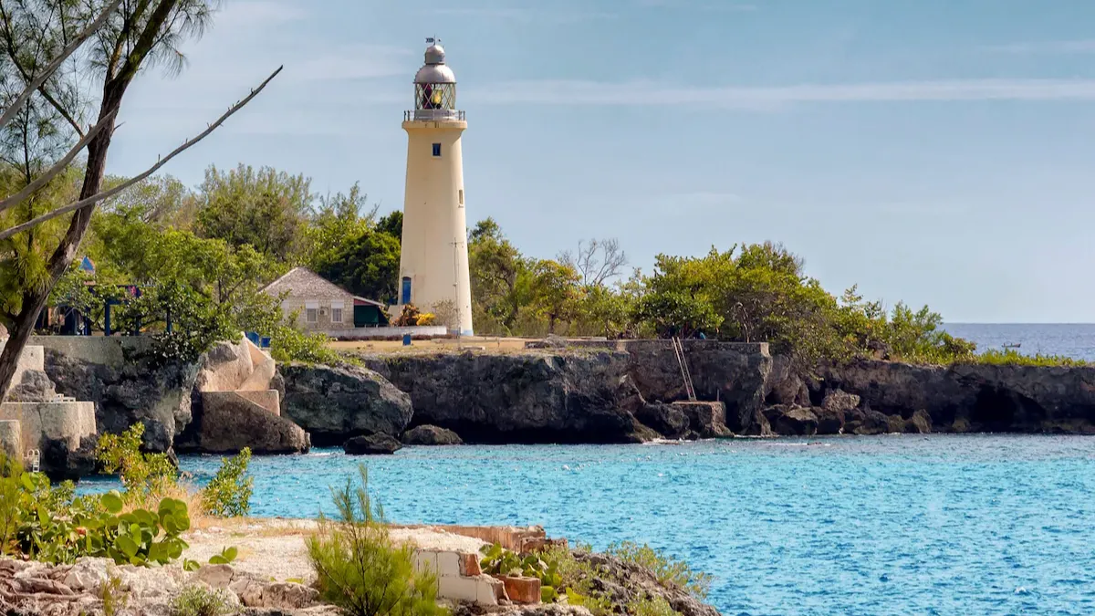 A white lighthouse stands on rocky shore by turquoise water, with green shrubs and a clear blue sky in a coastal scene.