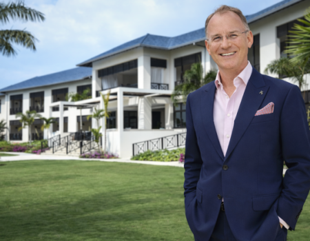 A well-dressed man in a navy suit stands smiling on a manicured lawn with white buildings and palm trees in the background.