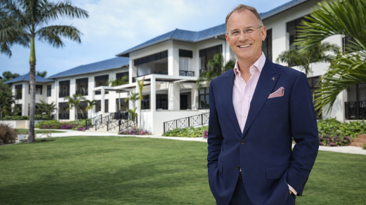 A well-dressed man in a navy suit stands smiling on a manicured lawn with white buildings and palm trees in the background.