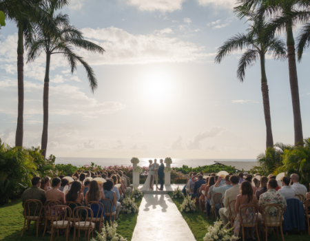 A tropical outdoor wedding ceremony on a sunny lawn, with palm trees, guests seated, a white aisle, and the ocean in the background, ending with the couple at the altar.