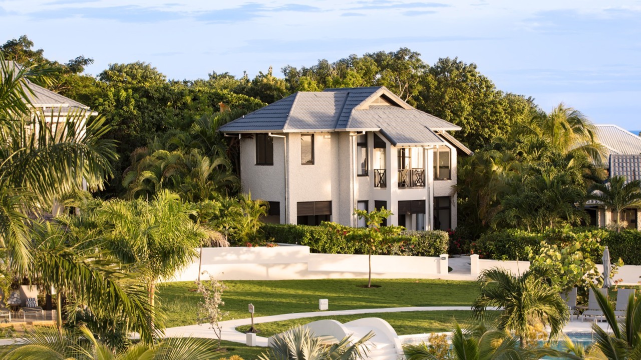 A modern two-story beige house with a gray roof, surrounded by palm trees and a white fence in a lush tropical setting.