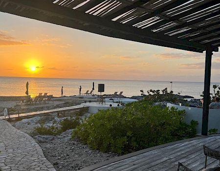 A sunset over a beach with a stone path, lounge chairs, and a shaded seating area in the foreground. The calm sea reflects warm orange light.