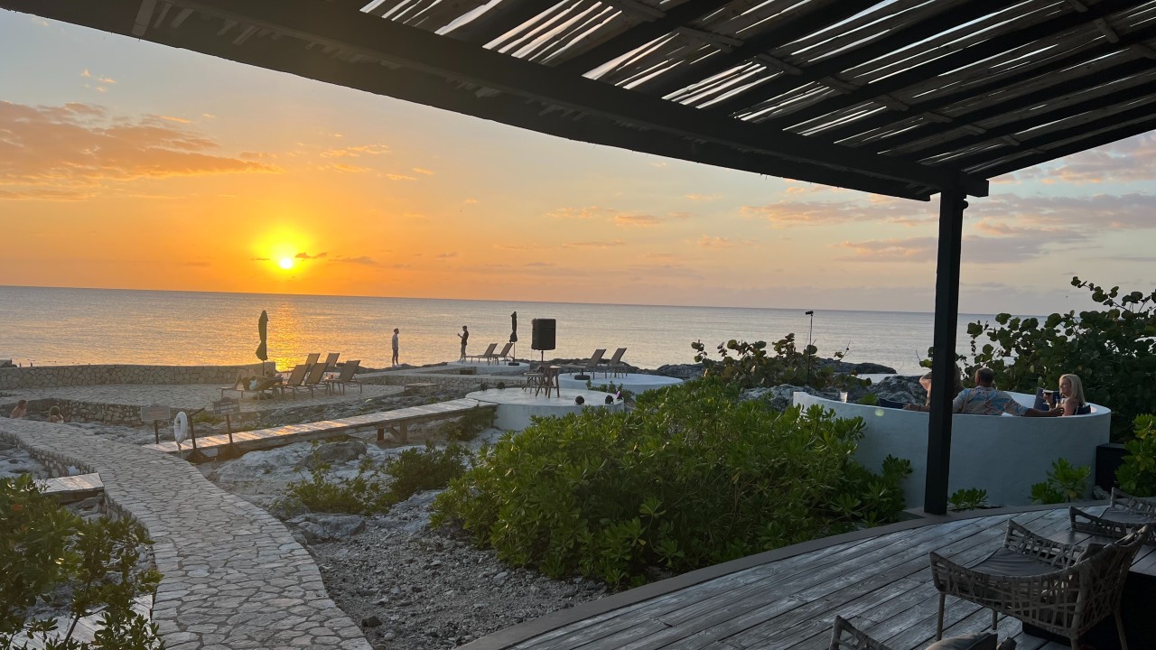 A sunset over a beach with a stone path, lounge chairs, and a shaded seating area in the foreground. The calm sea reflects warm orange light.