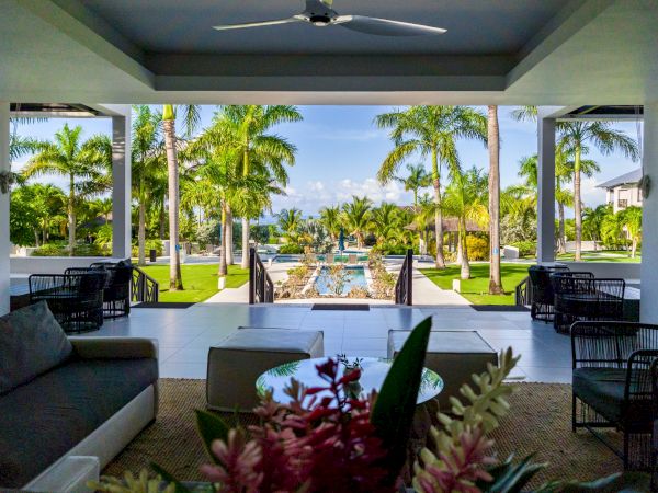 A luxurious outdoor patio overlooks a tropical resort pool with palm trees and modern seating under a shaded veranda, perfect for relaxing.